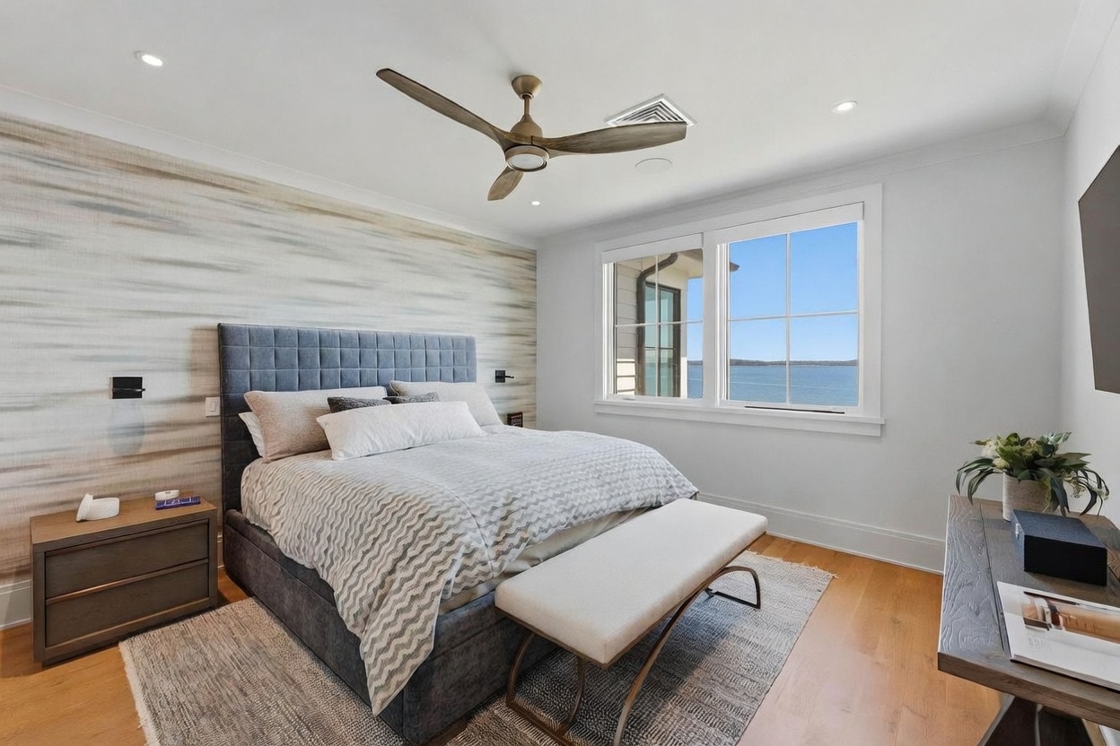This serene primary bedroom features a plush upholstered bed centered against a textured, neutral-toned accent wall. Large windows provide a clear, expansive view of the water, while a light wood bench sits at the foot of the bed atop a textured area rug. The room is completed by a modern ceiling fan and a sleek dresser, creating a calm and sophisticated atmosphere.