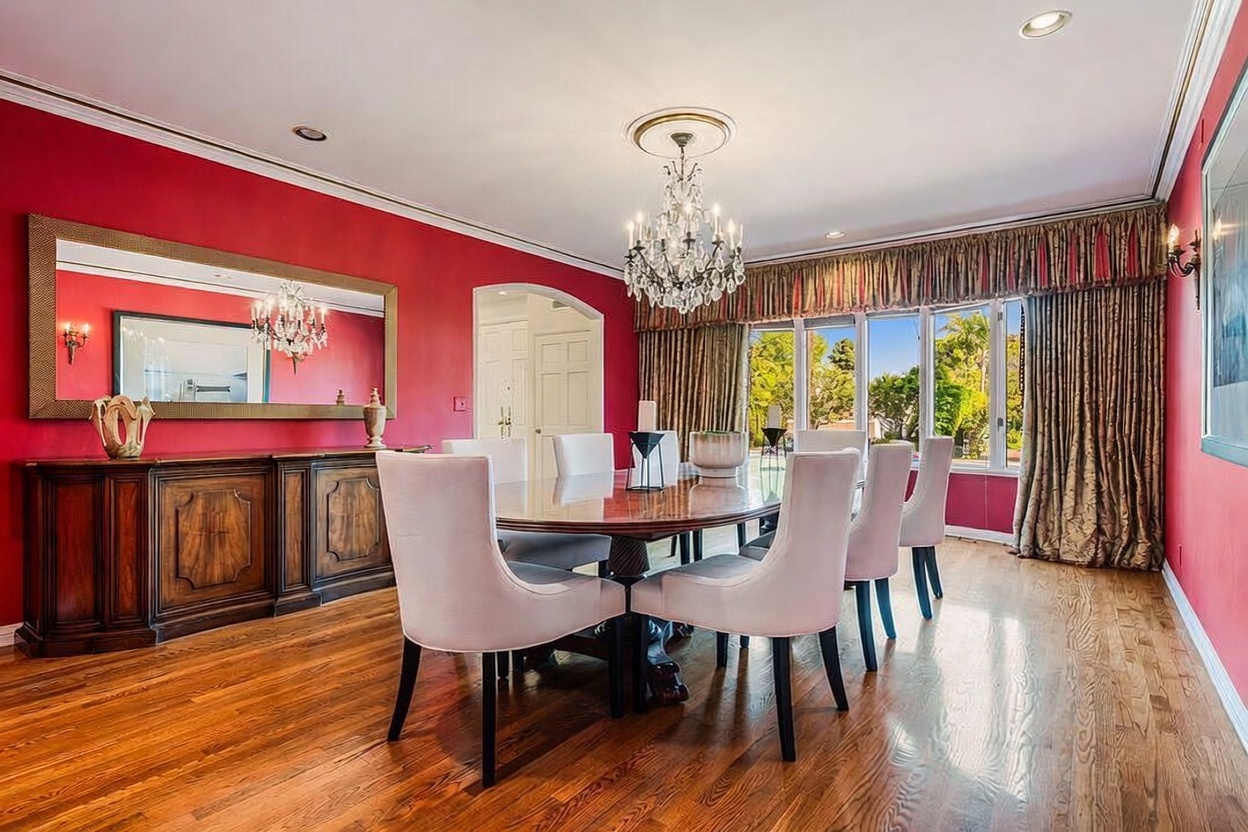 This formal dining room features bold red walls, rich hardwood flooring, and a large oval dining table surrounded by six upholstered chairs. A crystal chandelier serves as the centerpiece, hanging above the table, while a long wooden sideboard sits beneath a large, framed mirror on the wall. Large windows draped in patterned curtains allow natural light to fill the space, creating an elegant and traditional atmosphere.