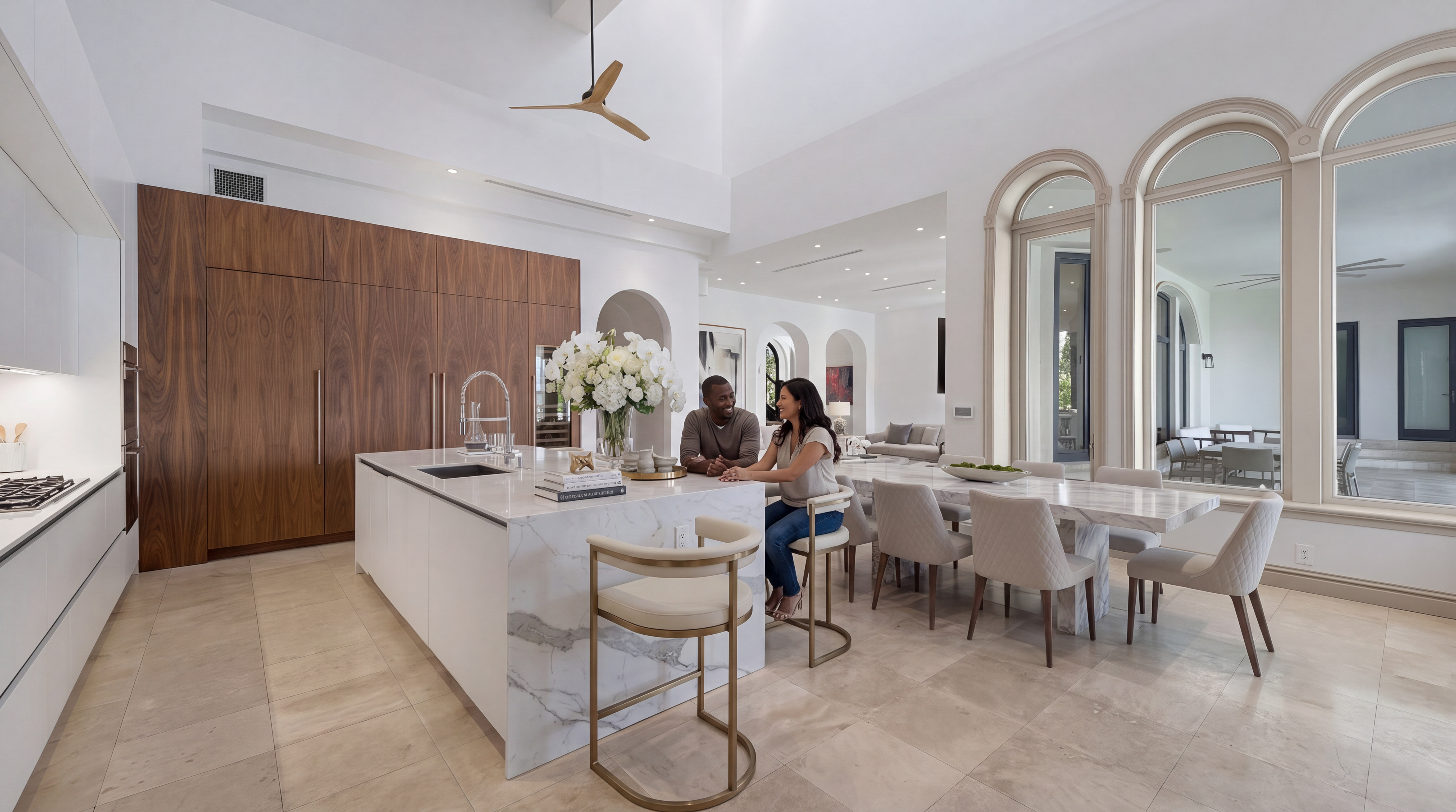 This stunning contemporary kitchen features a high-end design with seamless wood-paneled cabinetry and a large marble-topped island that serves as the centerpiece. The open-concept space includes a dedicated dining area and expansive arched windows that invite abundant natural light, creating an airy and sophisticated atmosphere. It is a perfect blend of luxury textures and modern aesthetic, ideal for both culinary tasks and social gatherings.