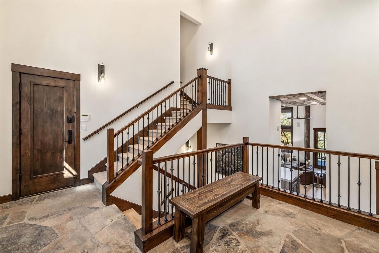 This grand entryway features a striking staircase with dark wood railings and iron balusters, set against crisp white walls. The flooring is composed of large, irregular stone tiles, and a rustic wooden bench sits at the base of the stairs, adding a warm, inviting touch. The perspective looks upward from the lower landing, highlighting the high ceilings and the transition into an adjacent living space visible through an open doorway.