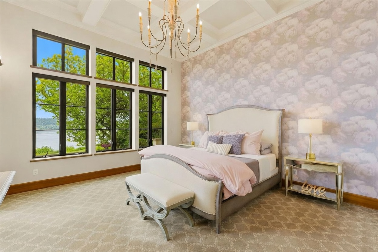 This elegant primary bedroom features a soft, romantic aesthetic with floral patterned wallpaper and a plush upholstered bed frame. Large windows provide an expansive view of the water and surrounding greenery, while a decorative chandelier hangs from the coffered ceiling. A matching bench sits at the foot of the bed, complemented by mirrored nightstands on either side.