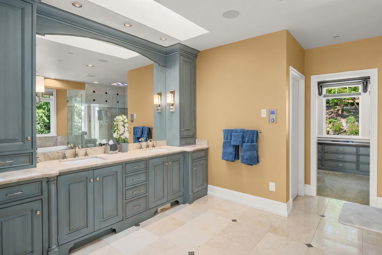 This elegant primary bathroom features a dual-sink vanity with custom blue-grey cabinetry and a large, arched mirror that enhances the sense of space. The room is finished with light-toned tile flooring, warm tan walls, and a seamless transition into a walk-in closet area. The overall aesthetic is sophisticated and well-appointed, offering a bright and airy atmosphere.