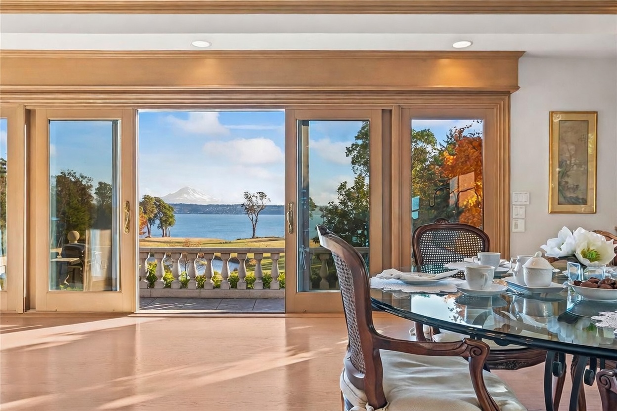 This dining area features a glass-topped table set for a meal, positioned in front of expansive sliding glass doors that frame a stunning view of a lake and a snow-capped mountain. The room is finished with light wood flooring and warm-toned wood trim around the large windows, creating a bright and inviting atmosphere. The perspective looks out from the dining space toward the outdoor balcony and the scenic landscape beyond.