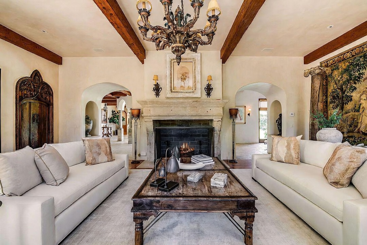 This elegant living room features a symmetrical layout with two white sofas facing a central wooden coffee table and a prominent stone fireplace. The space is defined by exposed wooden ceiling beams, a large ornate chandelier, and arched doorways leading to adjacent rooms. A decorative tapestry hangs on the right wall, contributing to the sophisticated, Mediterranean-inspired aesthetic of the home.