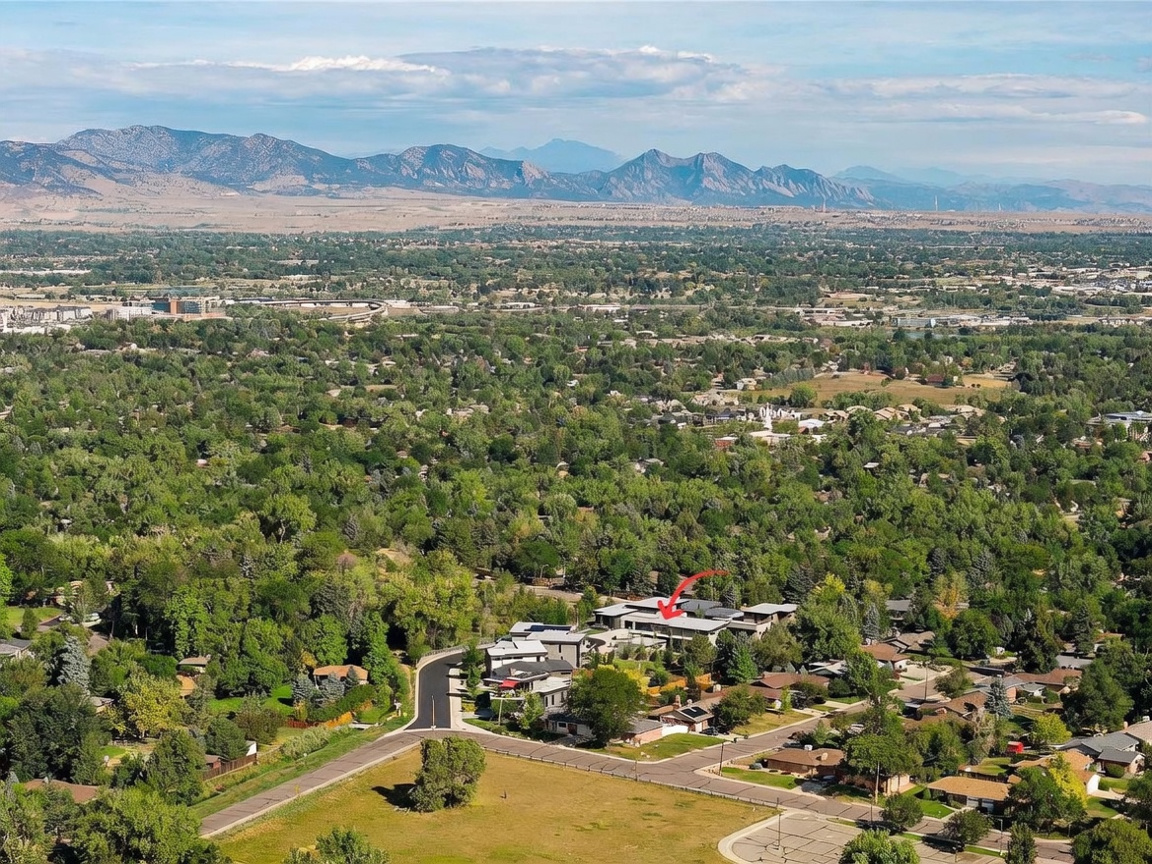 This high-altitude aerial view captures a modern residential complex nestled within a lush, tree-filled suburban neighborhood. A red arrow points toward the specific property, which features contemporary architecture with flat rooflines, while the background showcases a vast landscape leading to a prominent mountain range. The perspective provides a comprehensive look at the property's integration into the surrounding community and its scenic natural setting.