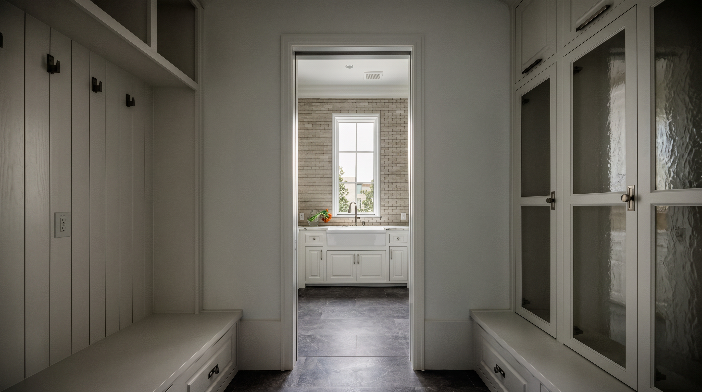 This view looks through a doorway into a bright laundry room featuring a deep farmhouse-style sink set into white cabinetry with a light-colored countertop. The room is framed by a mudroom area with built-in white storage lockers, bench seating, and glass-front cabinets on either side. The space is finished with dark, large-format floor tiles and a neutral color palette, creating a clean and organized transition between rooms.