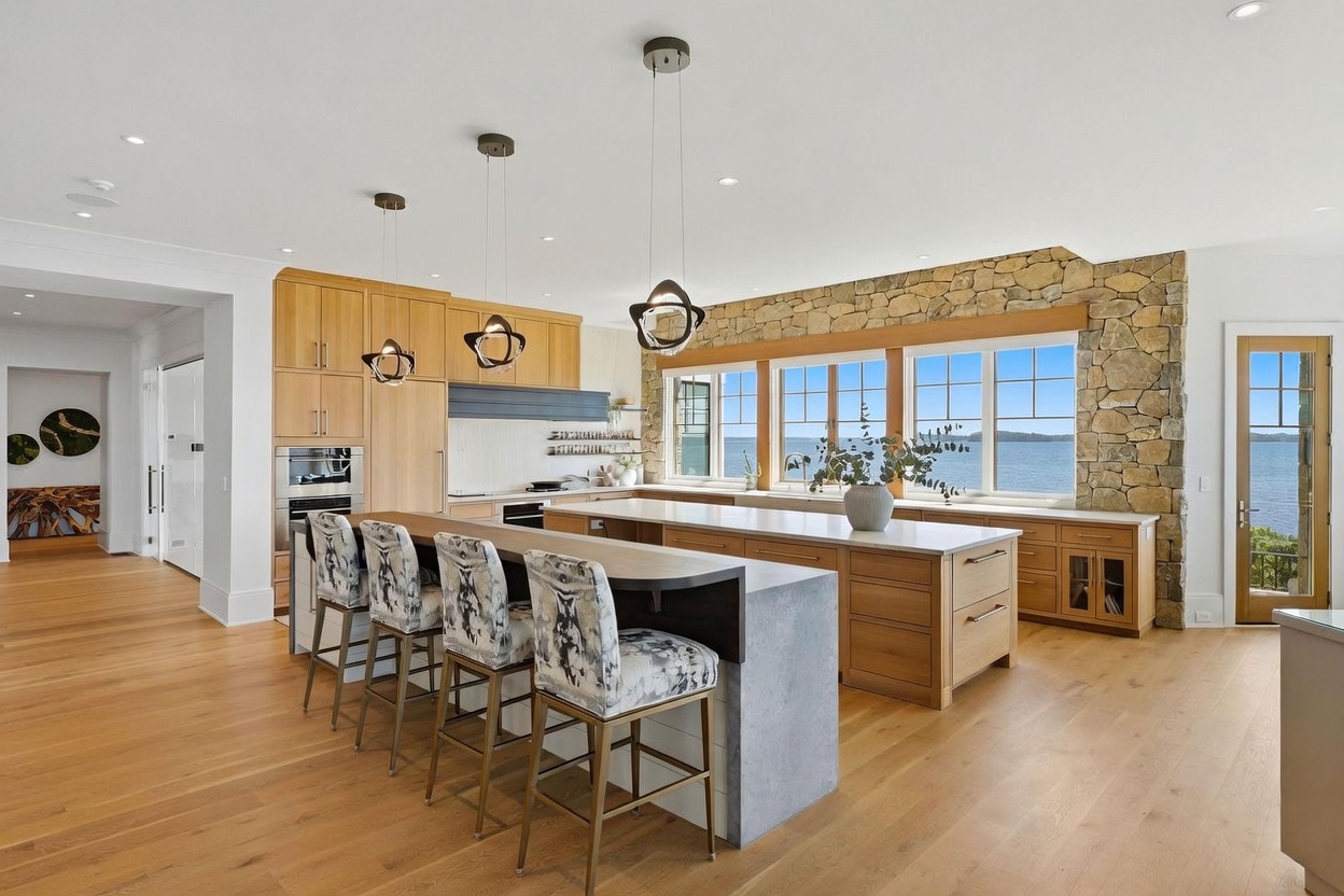 This bright, open-concept kitchen features light wood cabinetry, a large central island with bar seating, and a stone-clad wall surrounding the window area. The space is illuminated by modern pendant lighting and offers expansive views of the water through large windows. The overall aesthetic is warm and contemporary, emphasizing natural textures and a seamless connection to the outdoors.