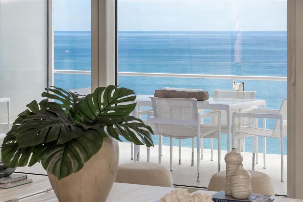 This interior view captures a dining area that seamlessly transitions to an outdoor balcony through large glass doors. In the foreground, a large potted plant with broad, split leaves sits on a light-toned table, while the background reveals a minimalist outdoor dining set overlooking the expansive ocean. The bright, airy atmosphere emphasizes the connection between the indoor living space and the coastal scenery.