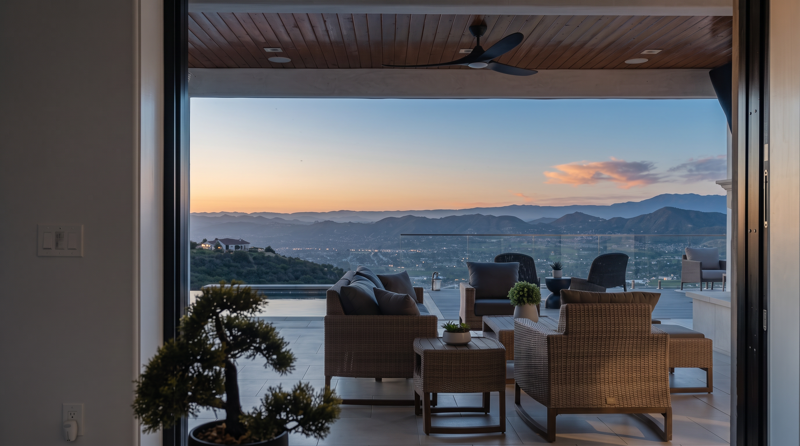 This stunning outdoor living space features a large covered patio with modern wicker furniture, providing an expansive panoramic view of the surrounding mountains and valley. The design seamlessly blends indoor and outdoor living, complemented by a sleek infinity-edge pool and sophisticated, clean-lined architecture. It is an ideal space for luxury entertaining and enjoying scenic vistas.