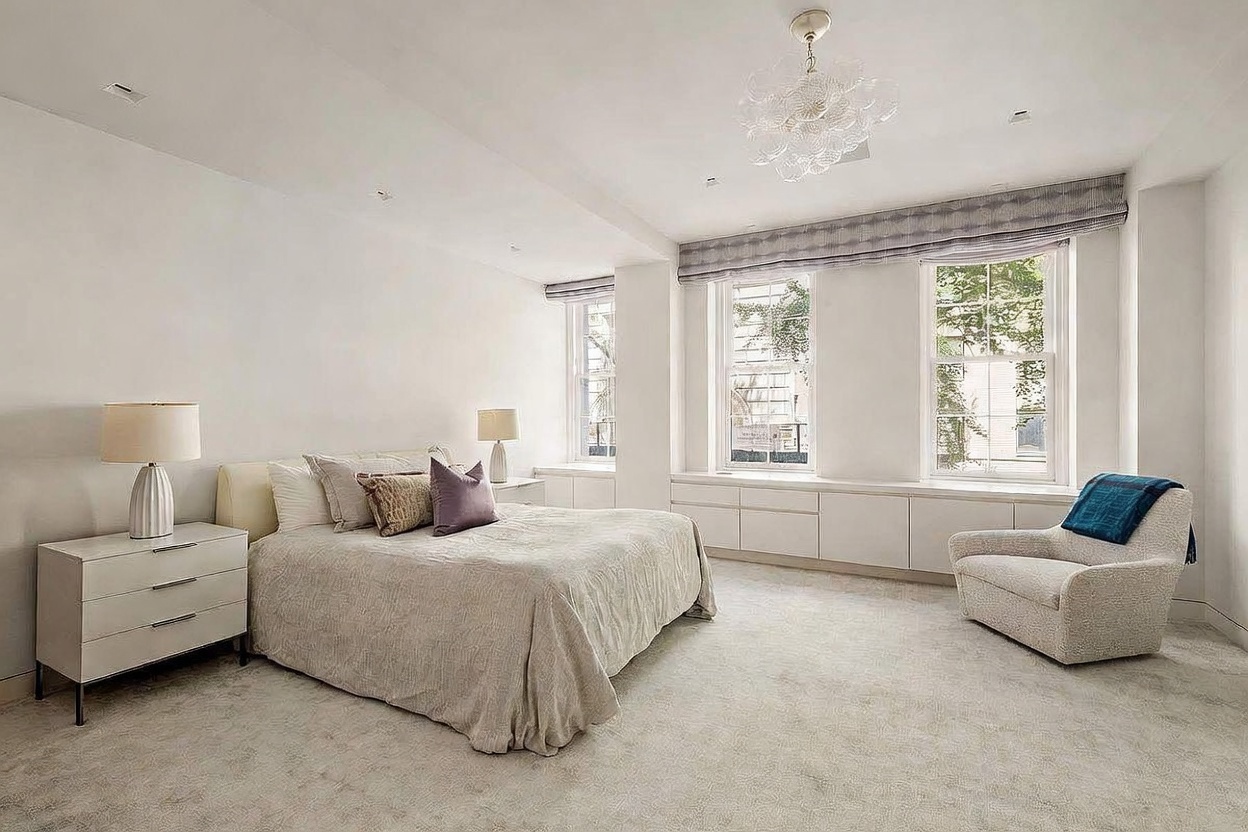 This spacious primary bedroom features a neutral color palette with soft carpeting and light-colored walls, creating a serene and airy atmosphere. The room is anchored by a large bed with layered bedding and decorative pillows, flanked by matching nightstands and modern table lamps. Large windows with tailored valances allow natural light to fill the space, while a comfortable armchair sits in the corner, providing a cozy seating area.