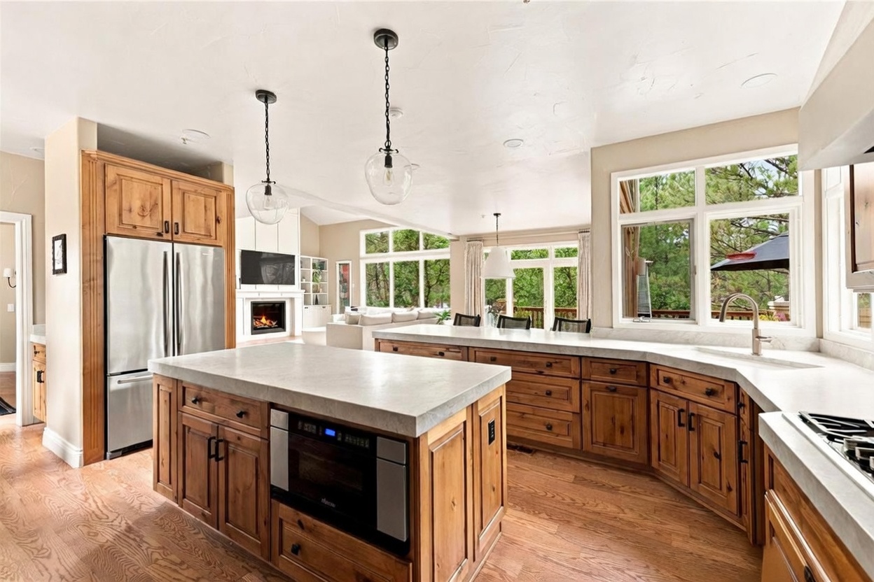 This spacious, open-concept kitchen features warm-toned wood cabinetry paired with light-colored stone countertops and a large central island. The space is illuminated by two glass pendant lights and flows seamlessly into a bright living area with a fireplace and large windows overlooking the trees. The perspective captures the functional layout of the kitchen, highlighting the stainless steel appliances and the inviting, airy atmosphere of the home.