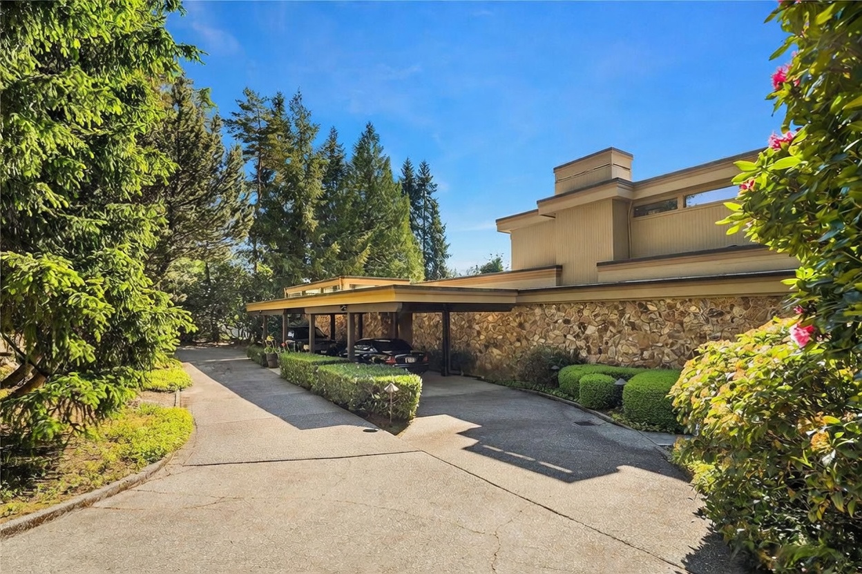 This exterior view captures a mid-century modern home featuring a prominent stone-clad facade and a long, flat-roofed carport. A concrete driveway leads toward the covered parking area, which is flanked by neatly manicured hedges and mature trees. The architectural style emphasizes horizontal lines and natural textures, creating a grounded and established aesthetic.