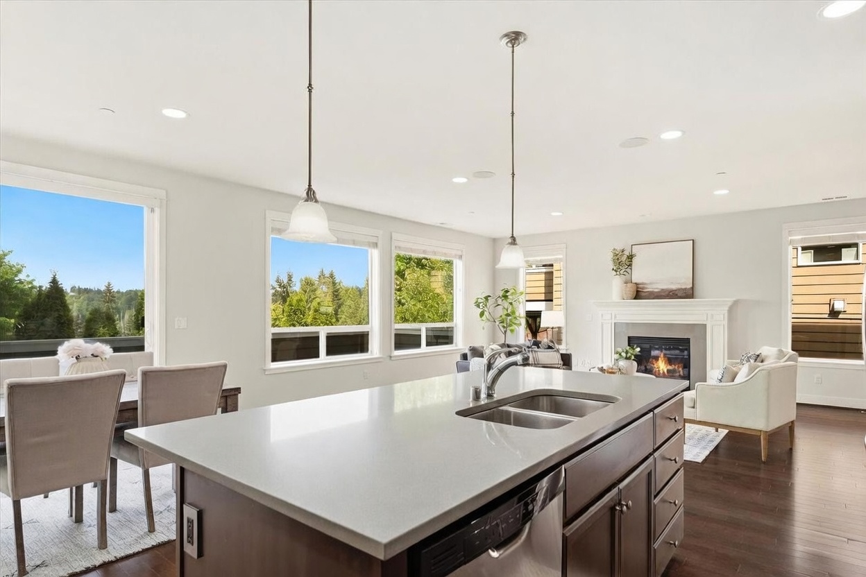This open-concept kitchen features a large central island with a double-basin sink and dark cabinetry, serving as the focal point of the space. Beyond the island, the room flows into a bright living area with a fireplace and comfortable seating, all illuminated by natural light from large windows. The space is finished with dark wood flooring and elegant pendant lighting, creating a warm and inviting atmosphere.