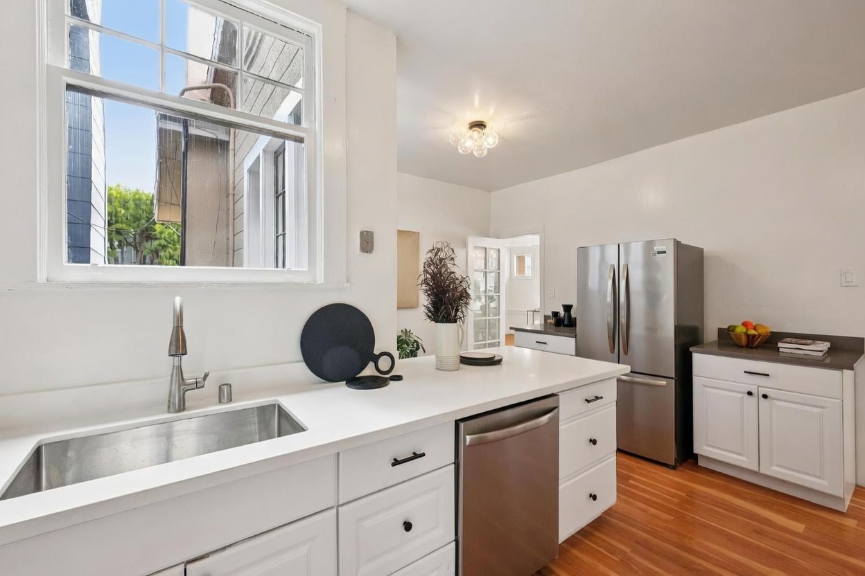 This bright, clean kitchen features white cabinetry paired with a white countertop and a stainless steel sink. A stainless steel dishwasher and refrigerator are integrated into the layout, which includes warm-toned wood flooring throughout. The space is illuminated by a modern multi-bulb ceiling fixture and natural light streaming through a large window above the sink.