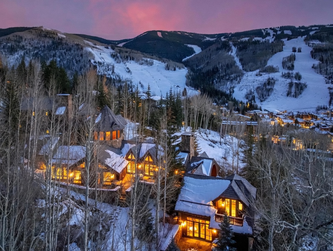This stunning aerial perspective captures a luxurious mountain estate nestled within a snowy, wooded landscape at twilight. The multi-level residence features rustic stone and timber architecture with warm, inviting interior lighting glowing through numerous windows, set against the dramatic backdrop of a ski resort and a vibrant pink-hued sunset. The composition highlights the home's integration into the rugged terrain, emphasizing its secluded yet accessible location near the slopes.