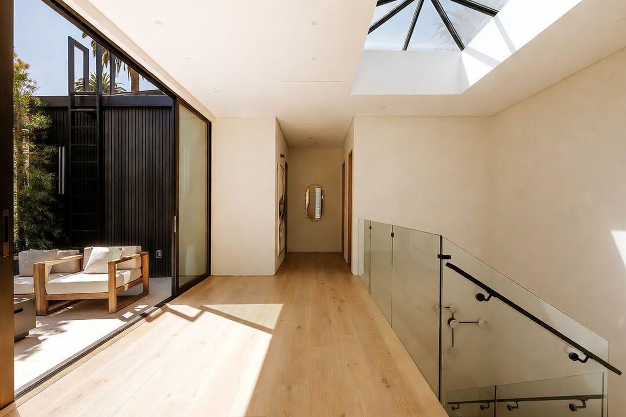 This bright, modern hallway features light wood flooring and a glass-paneled staircase railing that creates an open, airy feel. A large skylight above floods the space with natural light, while a sliding glass door opens to an adjacent outdoor patio area with lounge seating. The minimalist design is complemented by a sleek, oval mirror on the far wall, emphasizing a clean and sophisticated aesthetic.