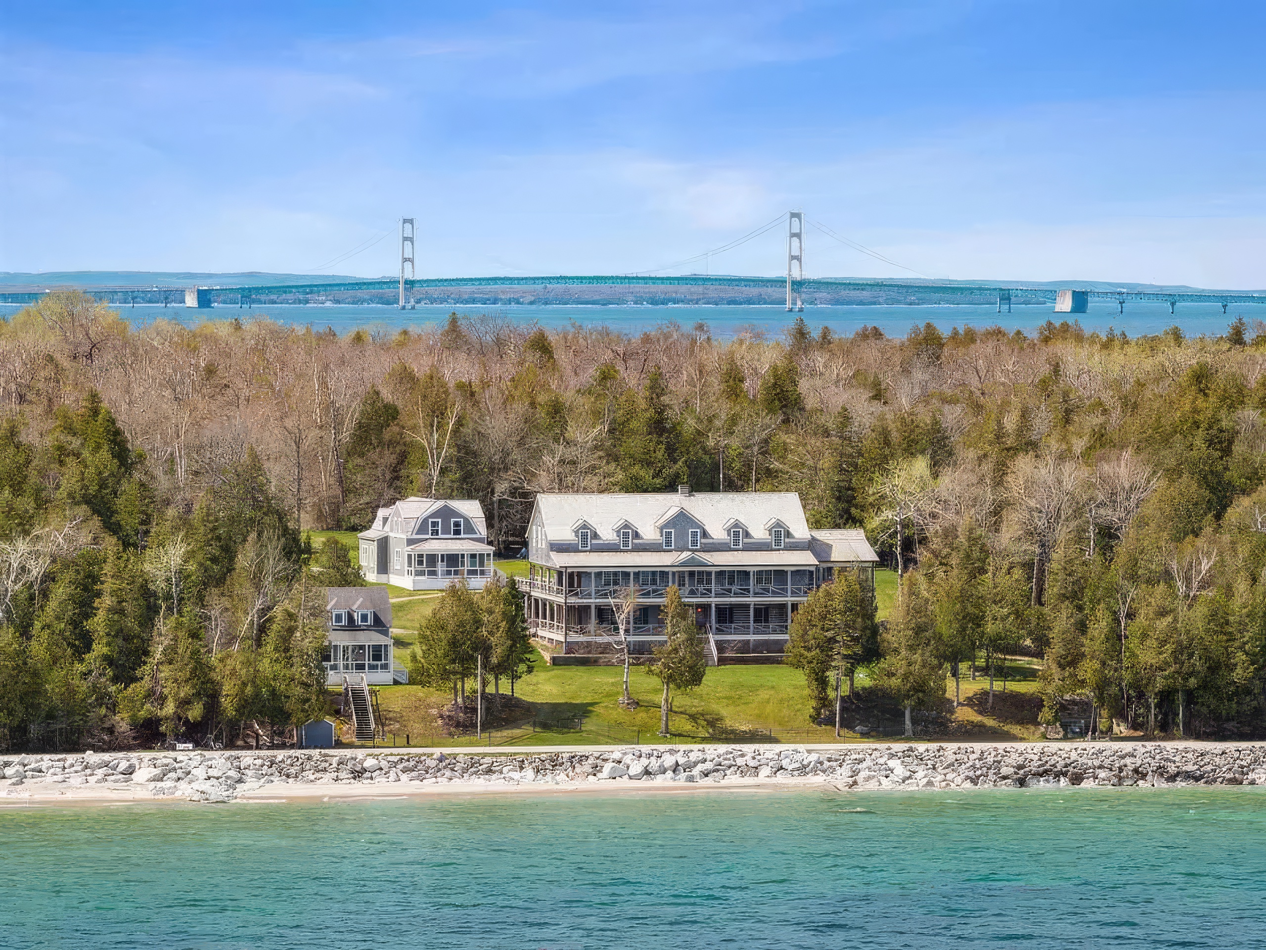 This aerial perspective captures a sprawling waterfront estate featuring a large, multi-story main residence and two smaller guest cottages nestled among dense, mature trees. The property sits directly on a rocky shoreline overlooking a vast body of water, with a prominent suspension bridge visible in the distance. The setting offers a serene, secluded atmosphere with expansive green lawns and direct access to the beach.