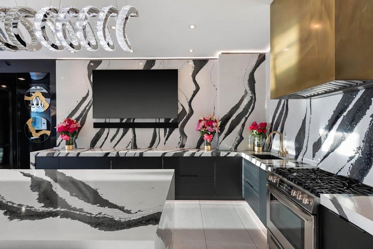 This high-end, contemporary kitchen features striking black-and-white veined stone surfaces on the countertops and backsplash, creating a bold, dramatic aesthetic. The space is equipped with dark cabinetry, a professional-grade gas range, and a prominent gold-toned range hood, while a large, modern crystal chandelier hangs above the foreground island. The perspective is a wide, eye-level shot that captures the seamless integration of luxury finishes and functional design.