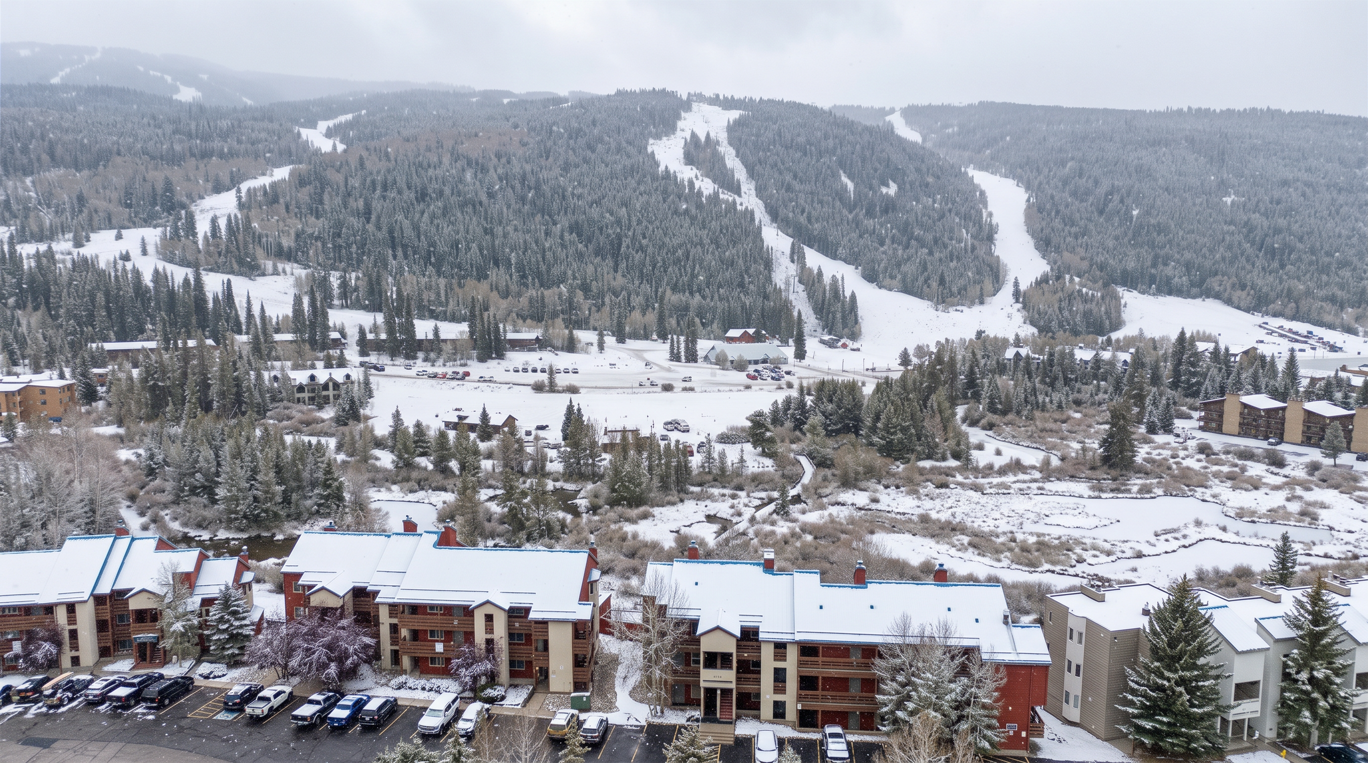 This aerial shot captures a charming mountain resort complex nestled in a snow-covered landscape during winter. The scene features multi-story timber-clad residential buildings with snow-dusted roofs, convenient surface parking, and a scenic backdrop of heavily forested slopes and ski trails. The environment suggests a peaceful, cold-weather retreat ideal for ski enthusiasts and nature lovers.