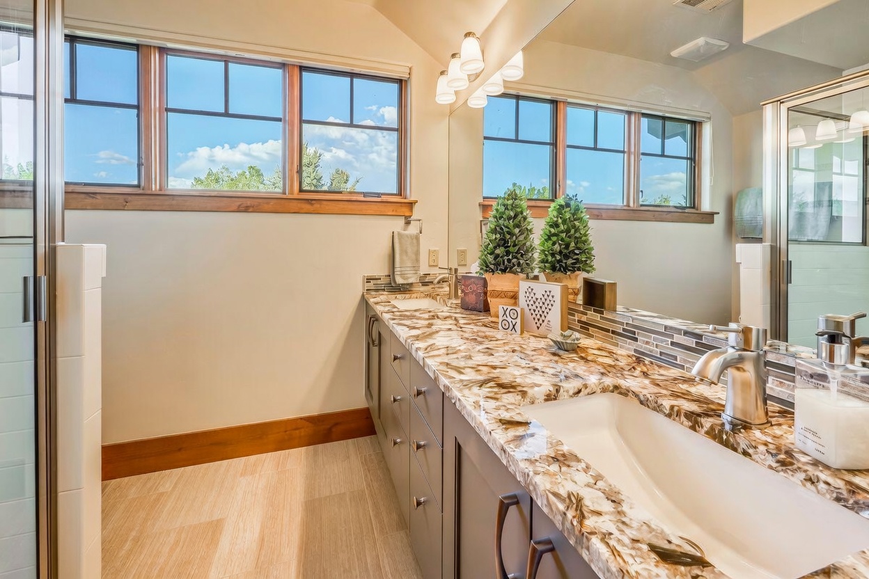 This bright and well-appointed bathroom features a long vanity with a striking, heavily veined stone countertop and dark gray cabinetry. A large mirror spans the wall above the vanity, reflecting the natural light from the windows and the decorative greenery placed on the counter. The space is finished with light-toned wood-look flooring and a glass-enclosed shower, creating a clean and modern aesthetic.