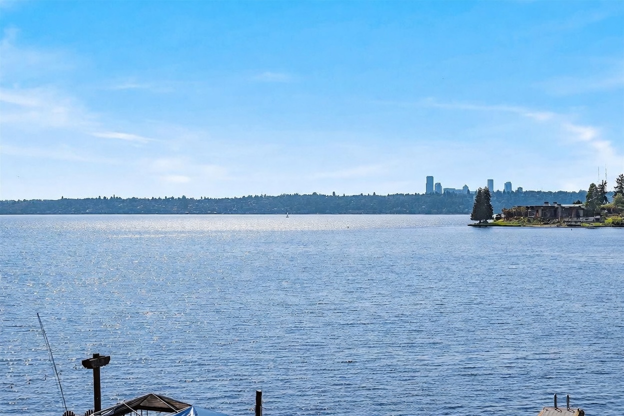 This wide-angle shot captures a serene waterfront view from an elevated perspective, likely a balcony or deck. The foreground features the tops of structures near the water, while the expansive lake stretches toward a distant shoreline marked by a city skyline. The bright blue sky and shimmering water create a peaceful, scenic atmosphere ideal for highlighting a prime waterfront location.
