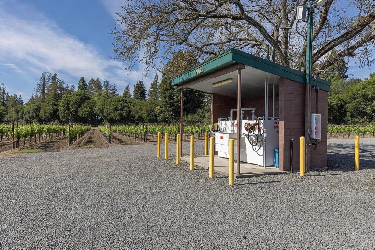 This image features a functional fuel station structure situated on a gravel lot adjacent to a vineyard. The structure is built with concrete blocks and a metal roof, housing fuel pumps and tanks protected by yellow safety bollards. The perspective is a wide-angle shot looking toward the vineyard, emphasizing the agricultural utility of the property.