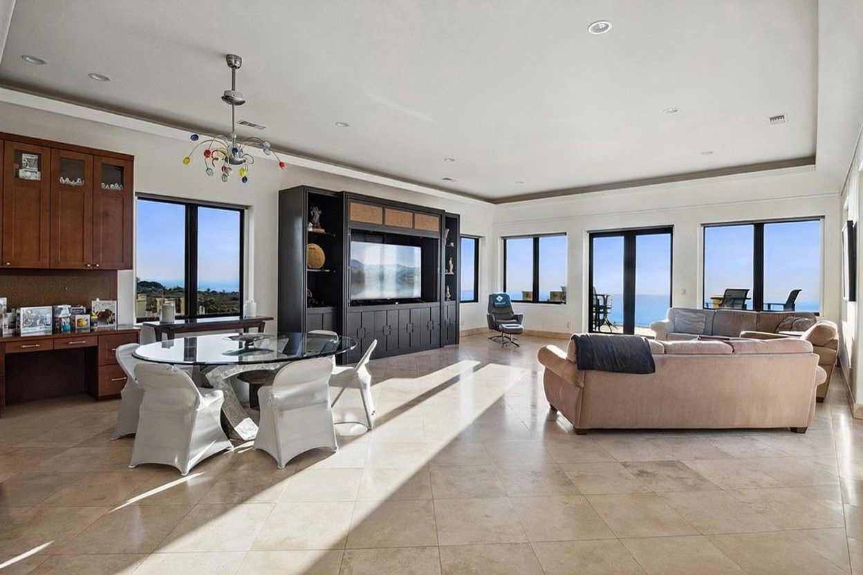 This spacious, open-concept living and dining area features large windows that provide expansive ocean views and abundant natural light. The room is anchored by a dark wood built-in media center and a matching desk unit, complemented by neutral tile flooring throughout. A glass-topped dining table sits in the foreground, while a comfortable seating area with a sofa and armchair occupies the space near the windows.