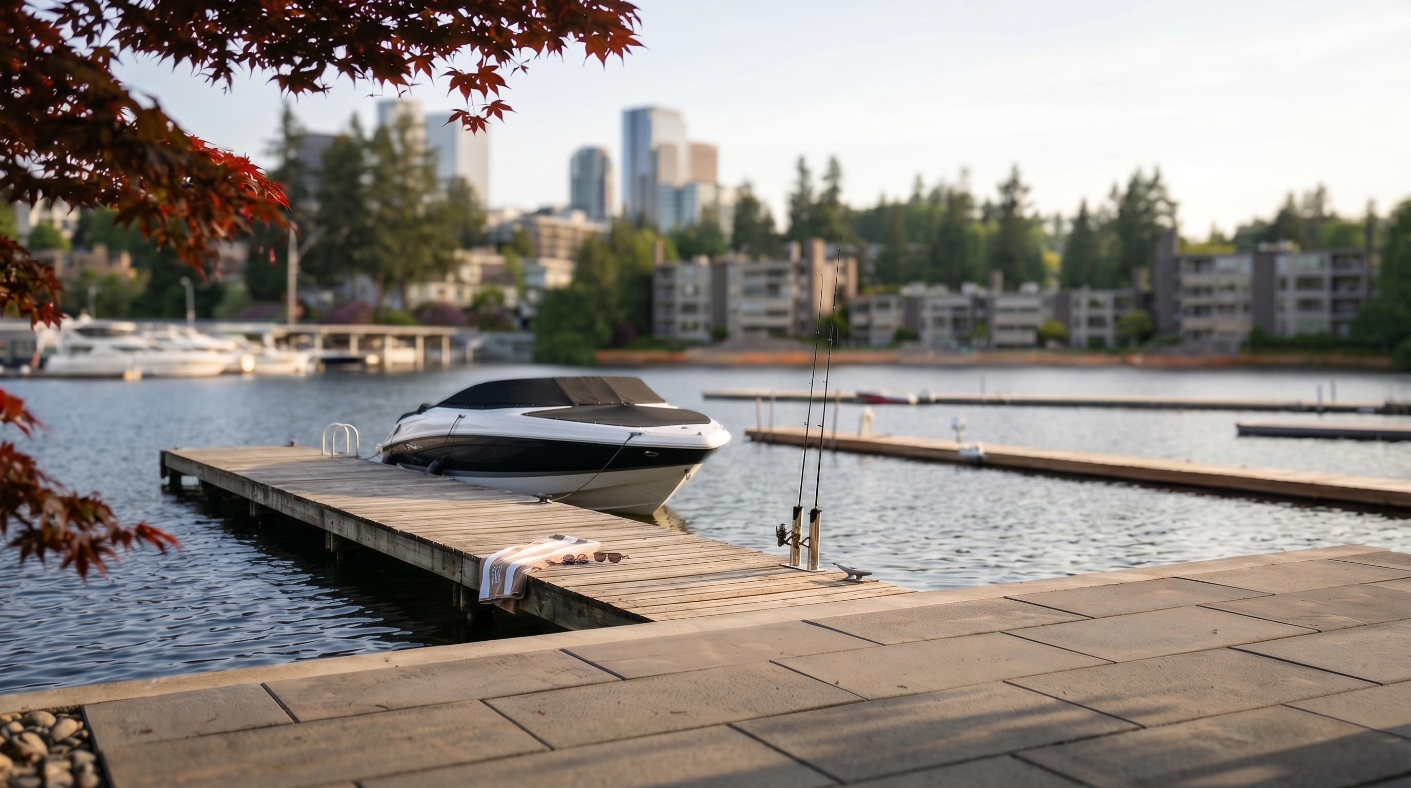This covered patio offers a serene, elevated view of a calm lake and a distant city skyline. The space is furnished with modern, light-toned wood-framed lounge chairs and features large glass doors that provide seamless indoor-outdoor flow. A metal railing lines the stairs leading down toward the private boat docks, creating a sophisticated and tranquil waterfront setting.