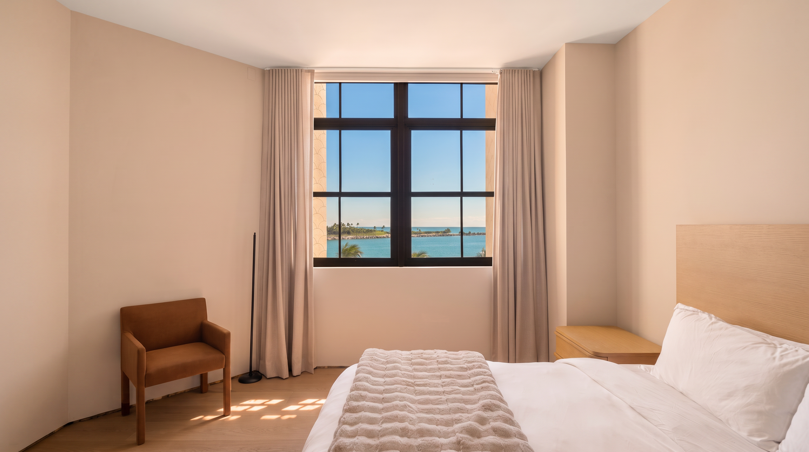 This bright and minimalist bedroom features a neutral color palette with light wood flooring and soft beige walls. A large window with floor-to-ceiling curtains frames a scenic view of the ocean, while a modern brown armchair sits in the corner. The room is furnished with a bed featuring a light wood headboard and a matching nightstand, creating a serene and airy atmosphere.
