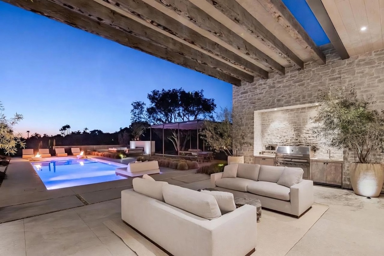 This luxurious outdoor living space features a covered patio area with a stone-clad wall housing a built-in grill and prep station. The space is furnished with comfortable light-toned sofas and overlooks a sleek, illuminated swimming pool set against a twilight sky. The architectural design incorporates exposed wooden beams overhead, creating a sophisticated and inviting atmosphere for evening relaxation.