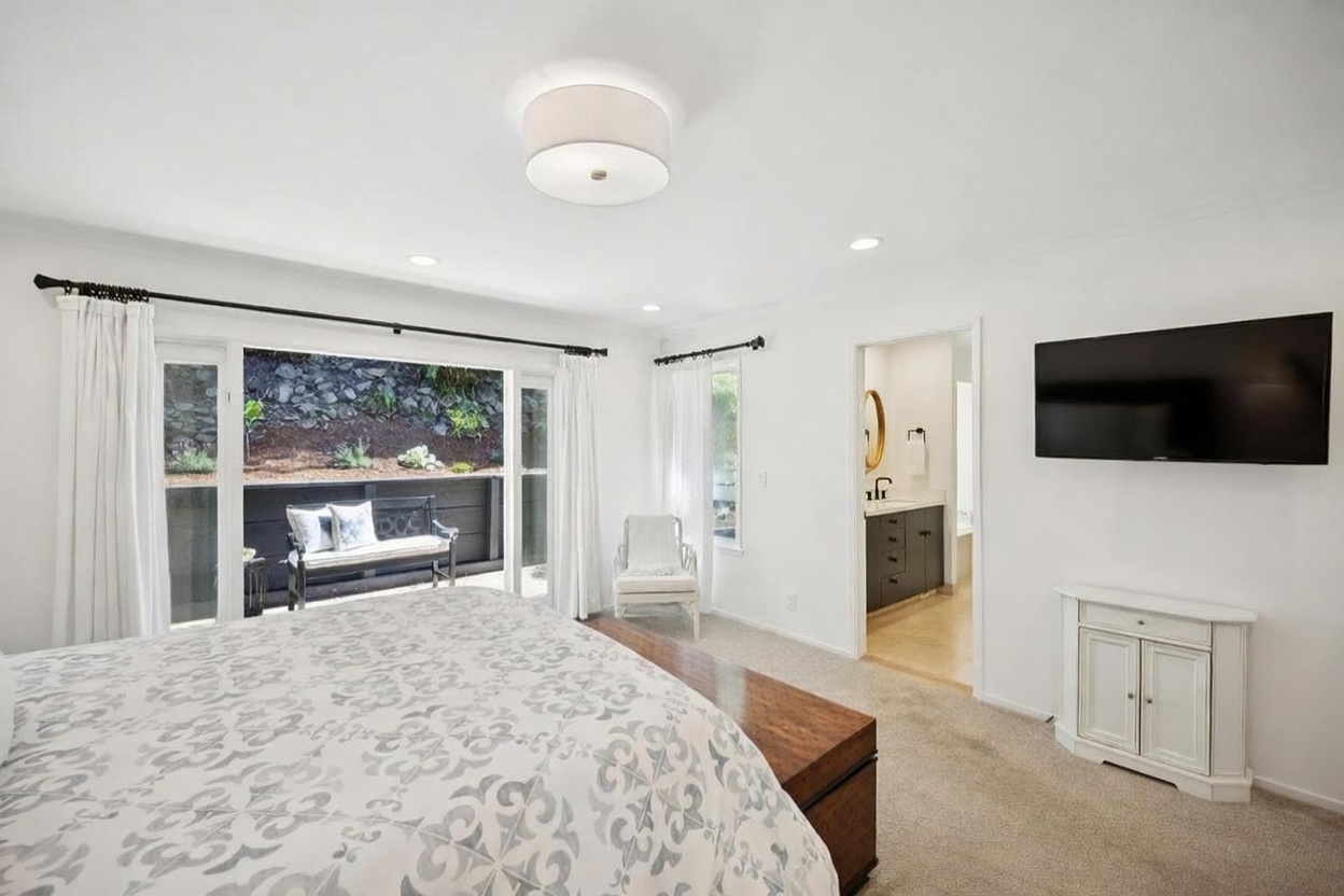 This bright and airy primary bedroom features a neutral color palette and a large sliding glass door that opens to a private outdoor patio area. The room is furnished with a patterned bedspread, a wooden storage chest at the foot of the bed, and a white media console beneath a wall-mounted television. An open doorway leads into an adjoining bathroom, creating a seamless flow between the sleeping and personal care spaces.