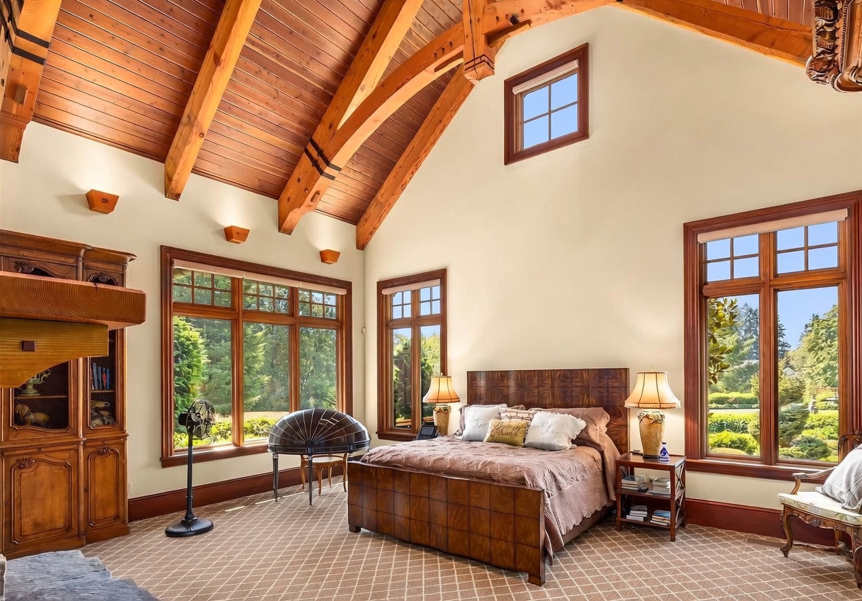 This spacious primary bedroom features a dramatic vaulted ceiling with exposed wooden beams and a warm wood-paneled finish. The room is anchored by a large bed with a dark wood headboard, flanked by matching nightstands and lamps, while large windows provide abundant natural light and views of the surrounding greenery. A patterned carpet covers the floor, and a unique, curved decorative piece sits near the window, contributing to the room's elegant and airy atmosphere.