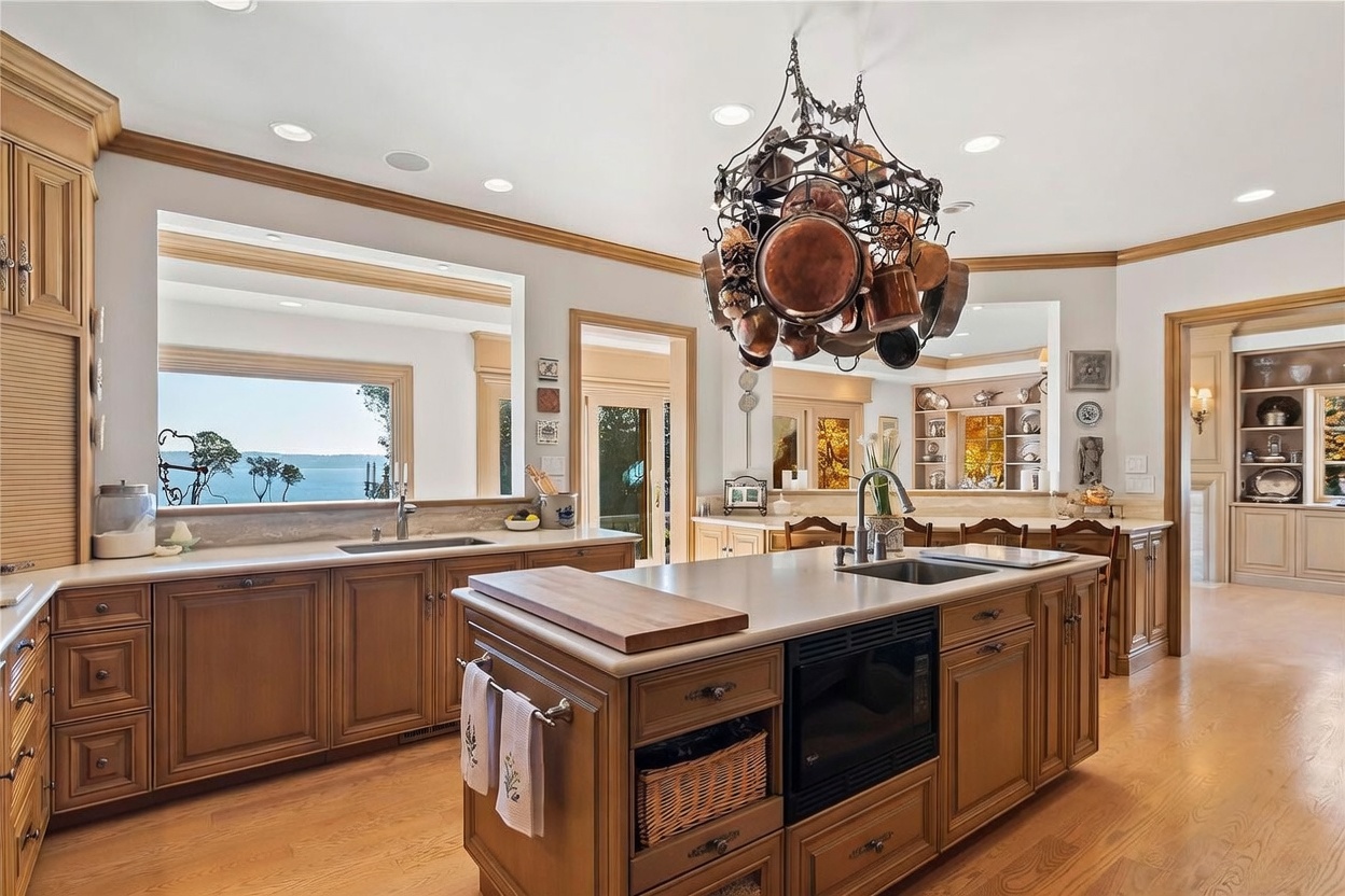 This spacious, traditional-style kitchen features warm-toned wood cabinetry, a large central island with a built-in microwave, and light-colored stone countertops. A prominent copper pot rack hangs above the island, while a large window over the sink provides a scenic view of the water. The room is well-lit with recessed lighting and offers a warm, inviting atmosphere with ample counter space for food preparation.