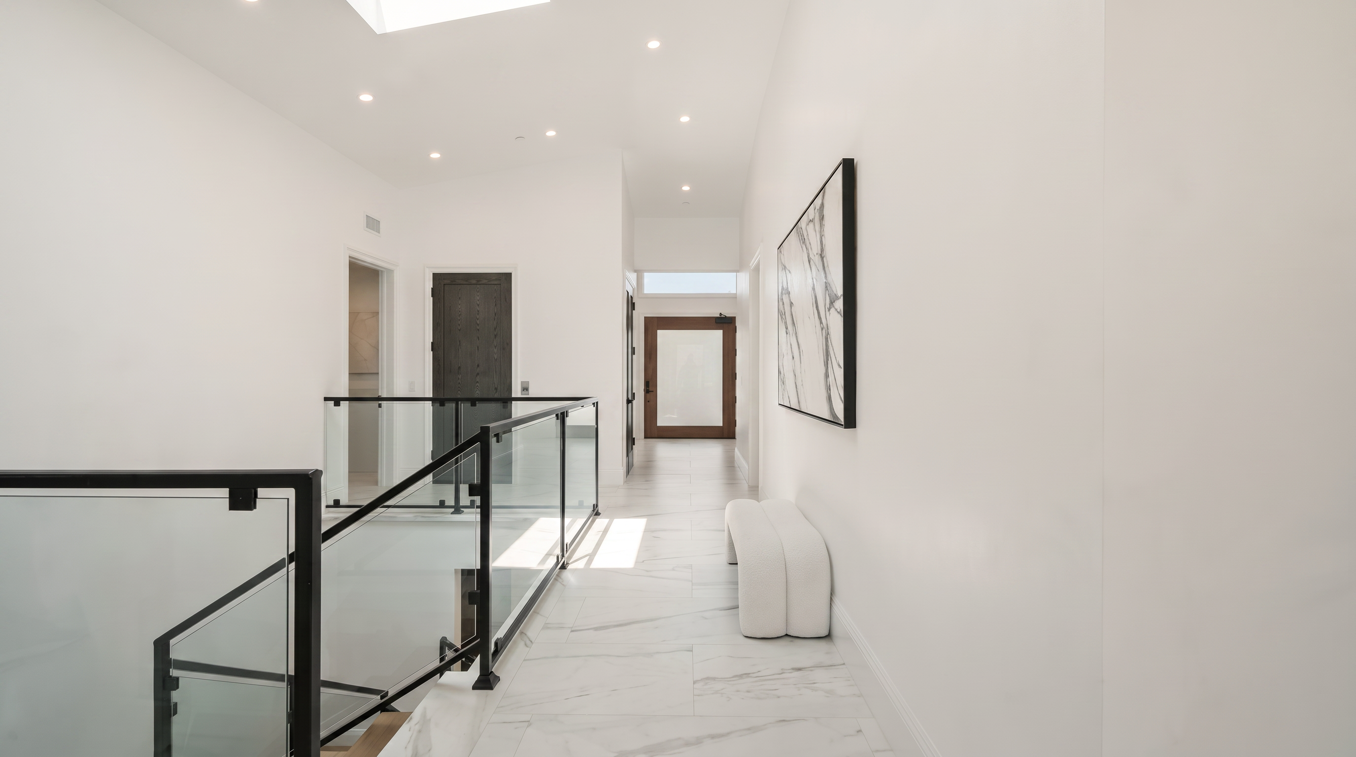 This bright, minimalist hallway features white walls and light-toned stone flooring with subtle veining. A glass railing with a dark frame overlooks the staircase, while a modern white bench and a large abstract art piece add a touch of sophistication to the space. The perspective looks down the corridor toward a doorway, emphasizing the clean lines and airy, open atmosphere of the home.