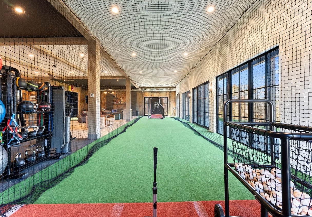 This expansive indoor sports training facility features a long stretch of artificial turf enclosed by protective netting, designed for batting or pitching practice. To the left, a dedicated weight training area is equipped with various kettlebells, medicine balls, and exercise machines, while the background reveals a lounge area with comfortable seating. The space is brightly lit by recessed ceiling lights and offers a clear, unobstructed view down the length of the training lane.