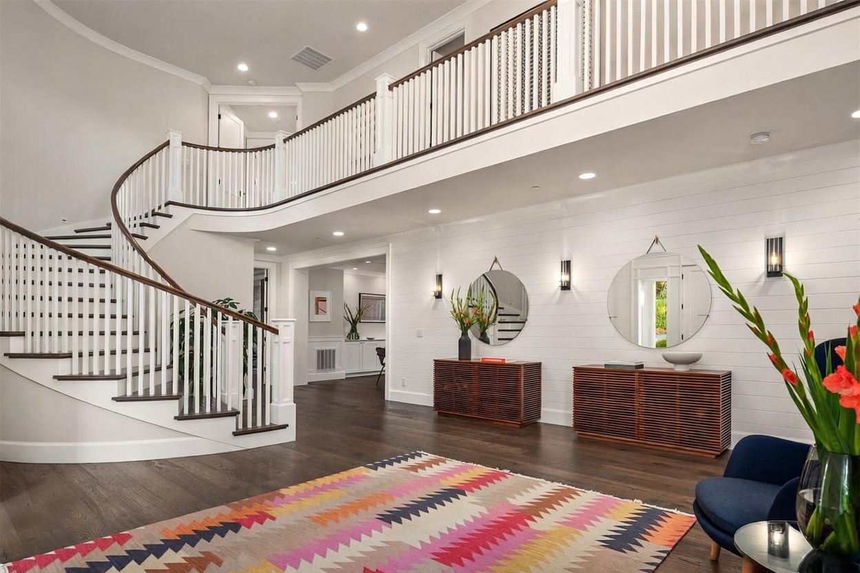 This grand entryway features a sweeping curved staircase with white railings and dark wood treads, creating a dramatic focal point. The space is finished with white shiplap walls, dark hardwood flooring, and a vibrant, multi-colored patterned rug that adds a modern touch. Two matching wooden consoles with slatted fronts are topped with circular mirrors and wall sconces, enhancing the bright and airy aesthetic of the foyer.