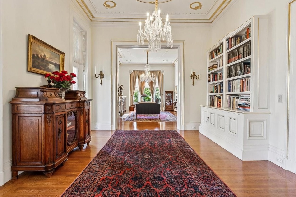 This elegant foyer features a long, ornate red patterned rug leading into a formal living area, framed by a grand doorway. To the left, a dark wood antique sideboard holds a vibrant floral arrangement, while a built-in white bookshelf lines the right wall. The space is illuminated by two matching crystal chandeliers, creating a sophisticated and traditional atmosphere.