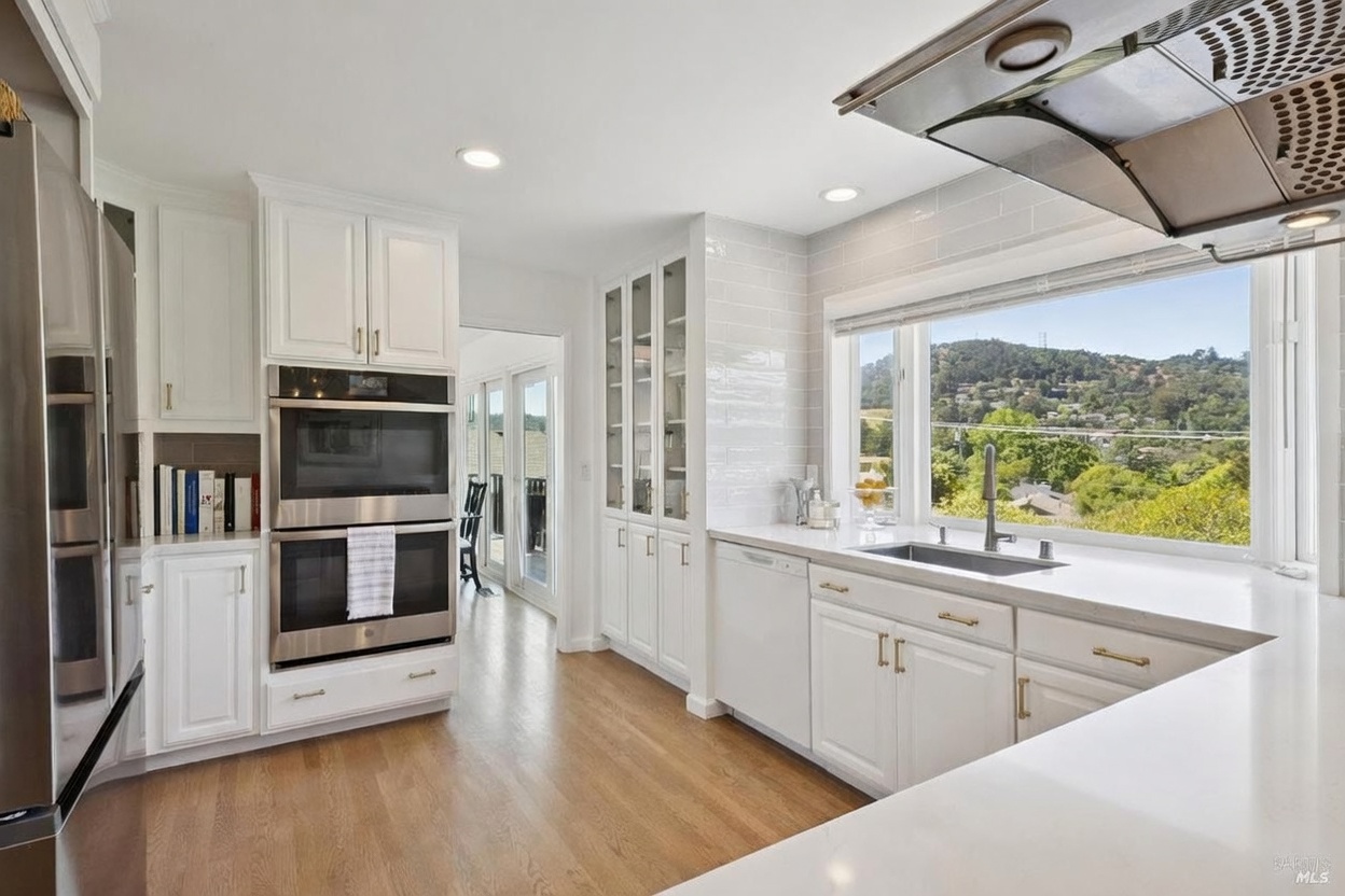 This bright and airy kitchen features white cabinetry, stainless steel appliances, and light-toned wood flooring. A large window above the sink provides a scenic view of the surrounding hills, while a range hood hangs above the counter space. The clean, modern aesthetic is complemented by neutral countertops and a classic subway tile backsplash.