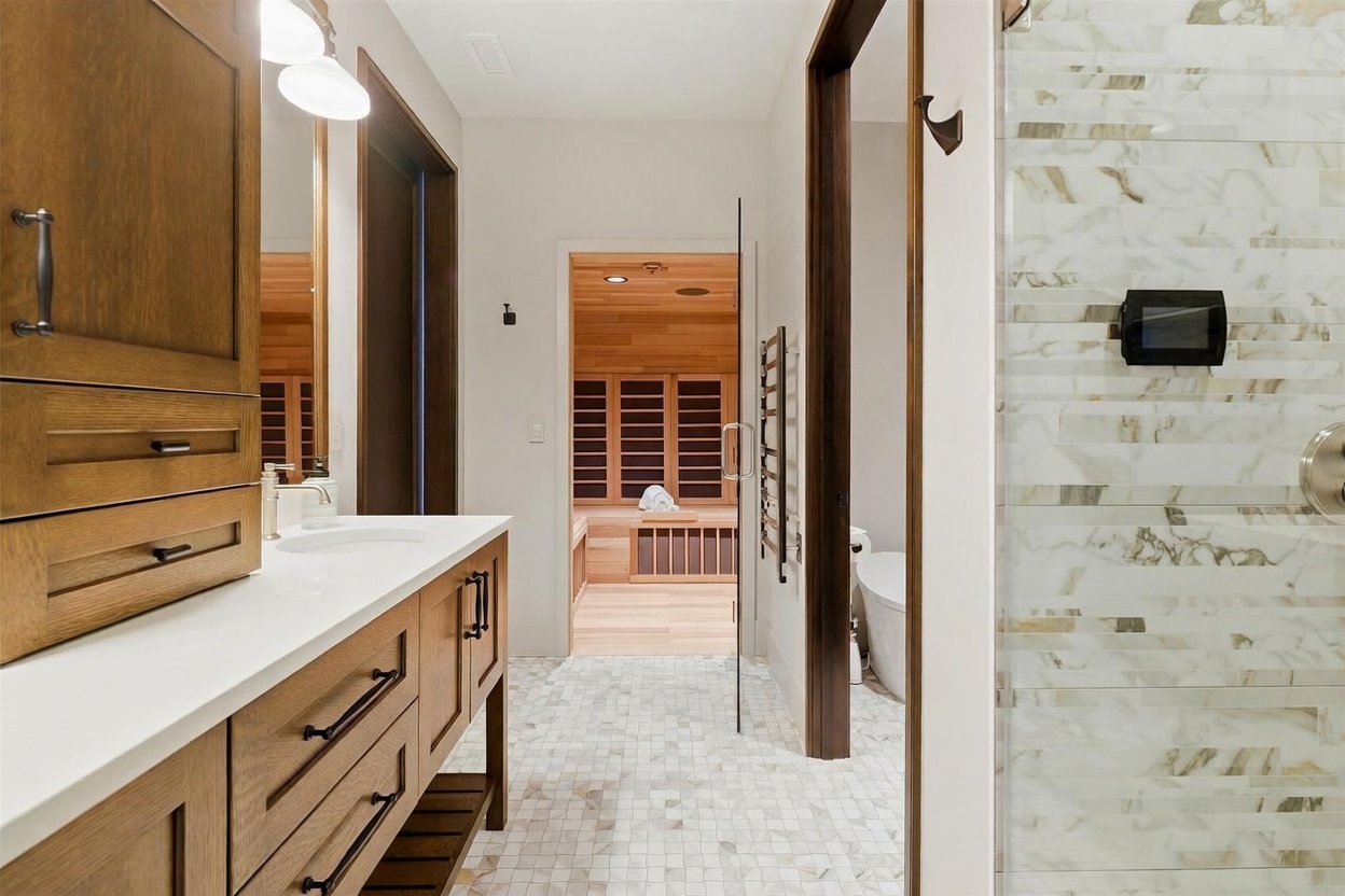 This primary bathroom features a warm wood vanity with a light-colored countertop and a glass-enclosed shower with patterned wall tile. A clear glass door leads into a dedicated sauna room, creating a spa-like atmosphere. The perspective looks through the bathroom toward the sauna, highlighting the clean lines and modern design of the space.