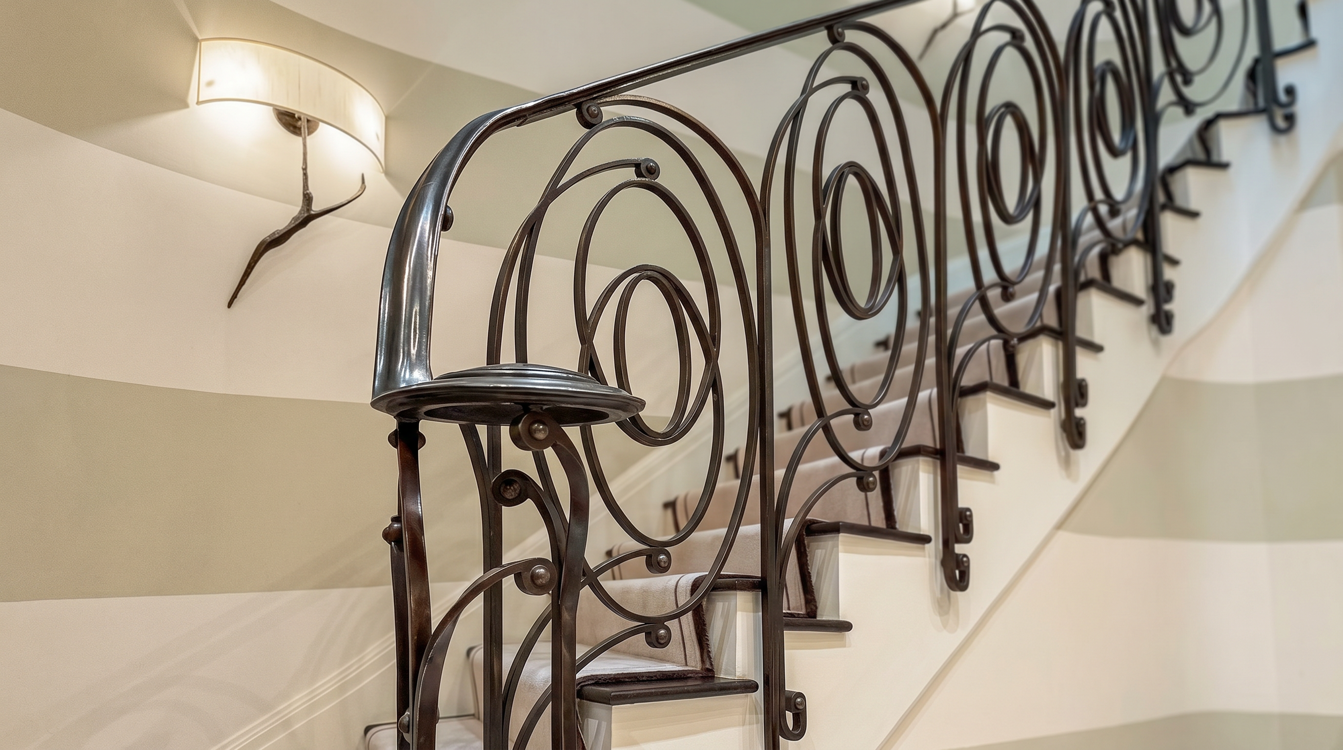 This elegant staircase features a striking wrought-iron railing with intricate, circular scrollwork patterns that create a sophisticated focal point. The steps are covered in a neutral-toned carpet with a dark border, complemented by walls painted in alternating horizontal stripes of cream and light beige. Two wall-mounted sconces with curved, organic-shaped arms provide warm, ambient lighting, enhancing the refined and classic aesthetic of the entryway.