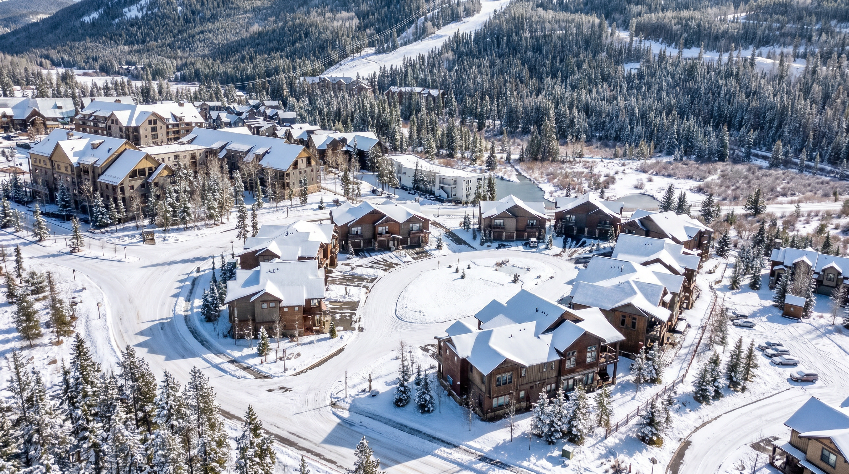 This high-angle aerial shot captures a sprawling mountain resort community nestled at the base of a vast, forested ski slope. The scene features a mix of multi-story residential buildings and townhomes with rustic, mountain-inspired architecture, surrounded by winding roads and natural landscape. The clear blue sky and expansive mountain backdrop create an inviting, scenic atmosphere perfect for a vacation destination.