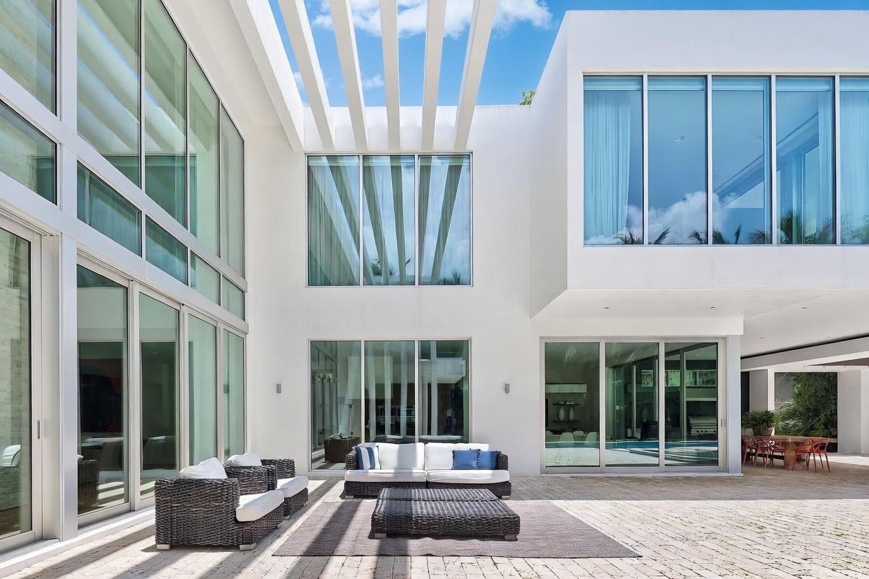 This modern, minimalist courtyard features a spacious stone-paved patio furnished with dark wicker outdoor seating and a matching ottoman. The area is framed by striking white architectural walls and expansive glass walls that provide a seamless transition between indoor and outdoor living spaces. Overhead, a series of white beams creates a pergola-like structure, casting linear shadows across the patio floor.