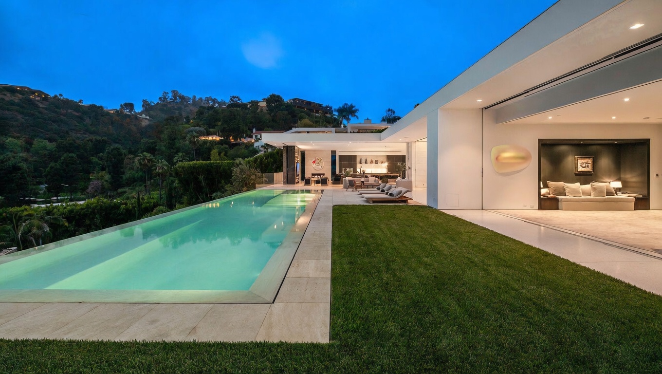 This stunning modern luxury estate features a sleek, long infinity-edge pool that seamlessly blends with the lush hillside landscape at dusk. The expansive outdoor living area includes a manicured lawn and a covered patio space that transitions effortlessly into a sophisticated, minimalist interior living room. The architectural design emphasizes clean lines, open-concept flow, and a seamless connection between indoor and outdoor environments.