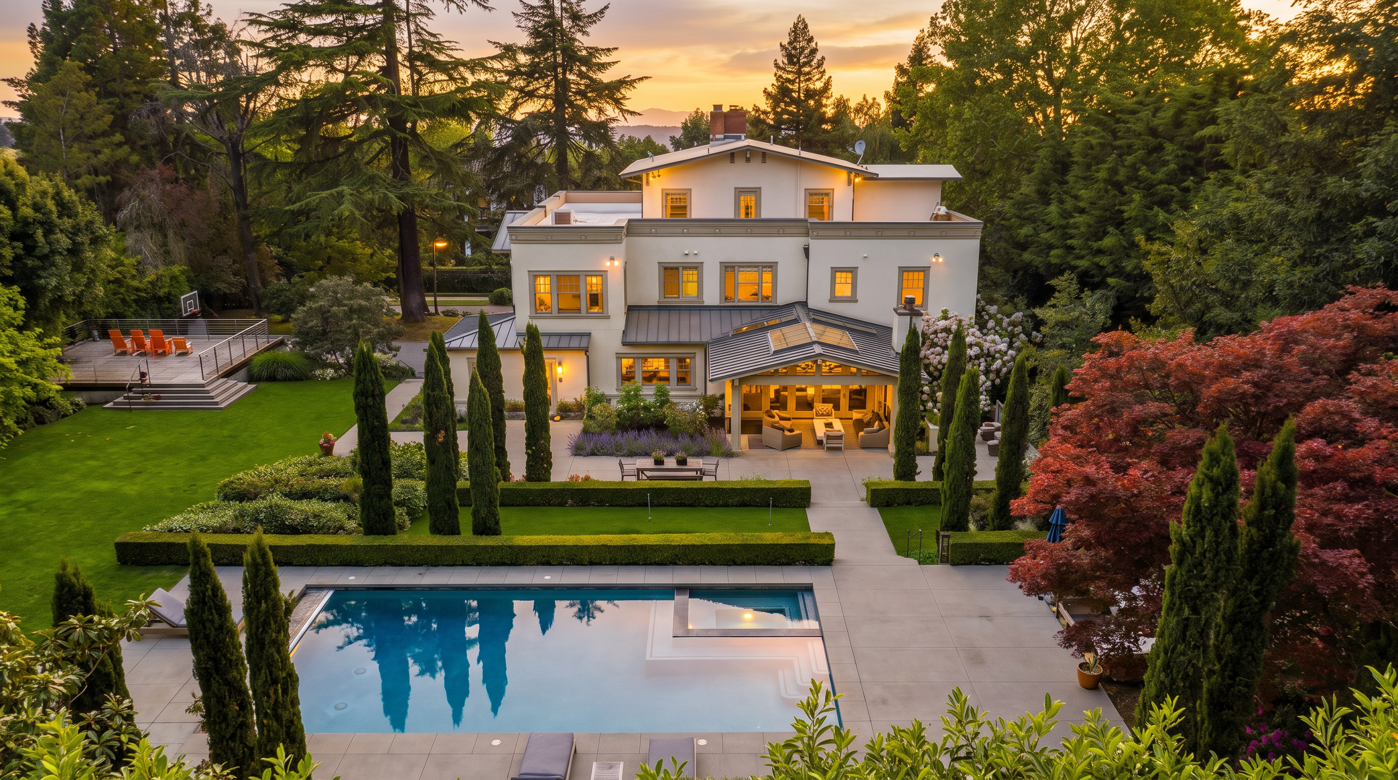 This high-angle aerial view captures a grand, multi-story estate set within a lush, landscaped property. The backyard features a large rectangular swimming pool with an integrated spa, a manicured lawn, a covered patio area, and a separate elevated deck, all surrounded by mature trees and structured hedges. The home showcases a classic architectural style with a light-colored facade, a dark roof, and a covered outdoor living space, creating a sophisticated and private retreat.