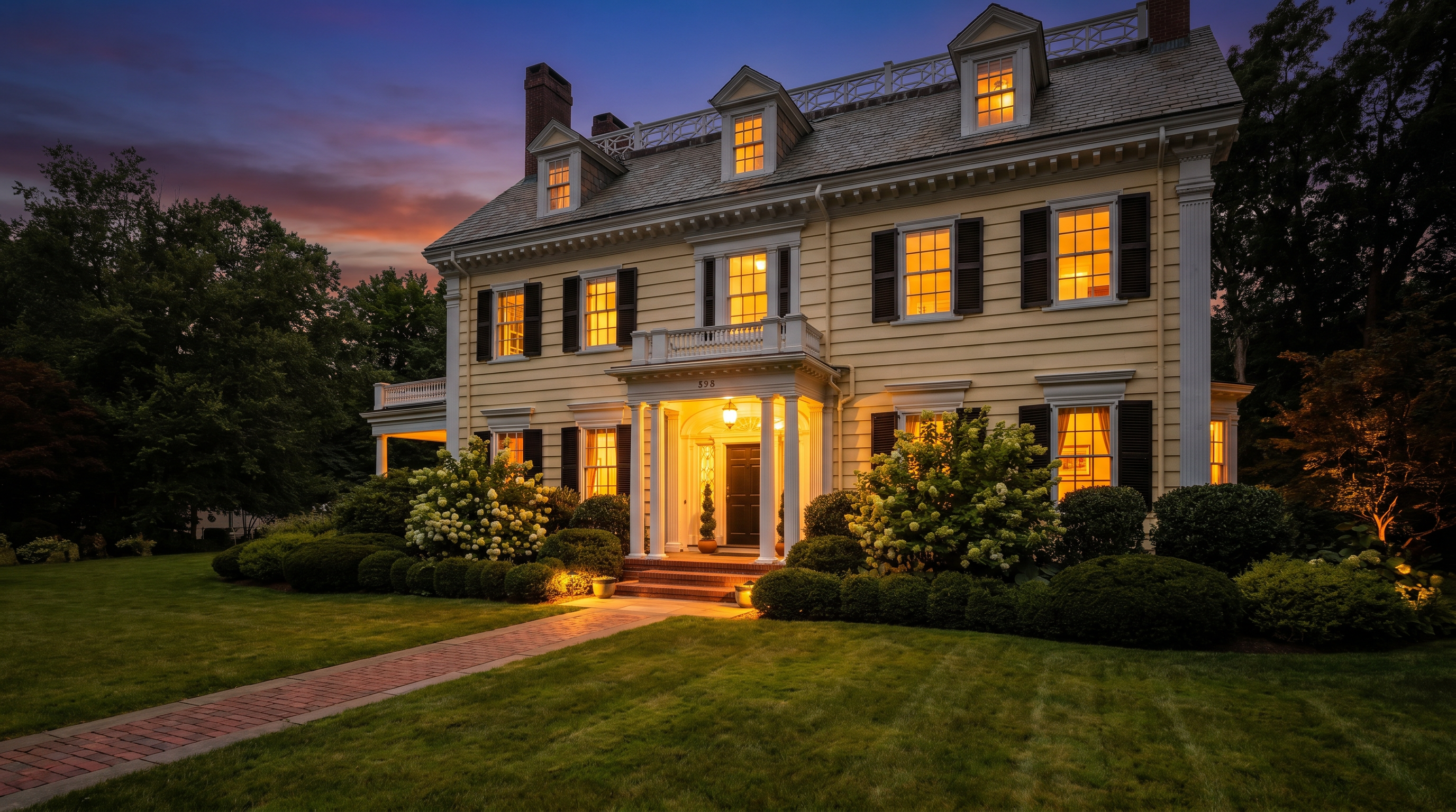 This classic, yellow-sided colonial home features a symmetrical facade with a prominent covered entryway supported by white columns. A brick walkway leads through a manicured lawn toward the front door, framed by lush green shrubs and blooming hydrangea bushes. The perspective captures the home's stately curb appeal and well-maintained landscaping under a soft, overcast sky.