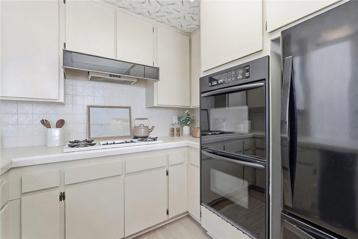 This kitchen features light-colored cabinetry paired with a white tiled backsplash and a white countertop. A built-in double oven is integrated into the cabinetry alongside a black refrigerator, while a gas cooktop sits beneath a range hood. The space is accented with a framed piece of art and a small kettle, creating a functional and compact culinary area.