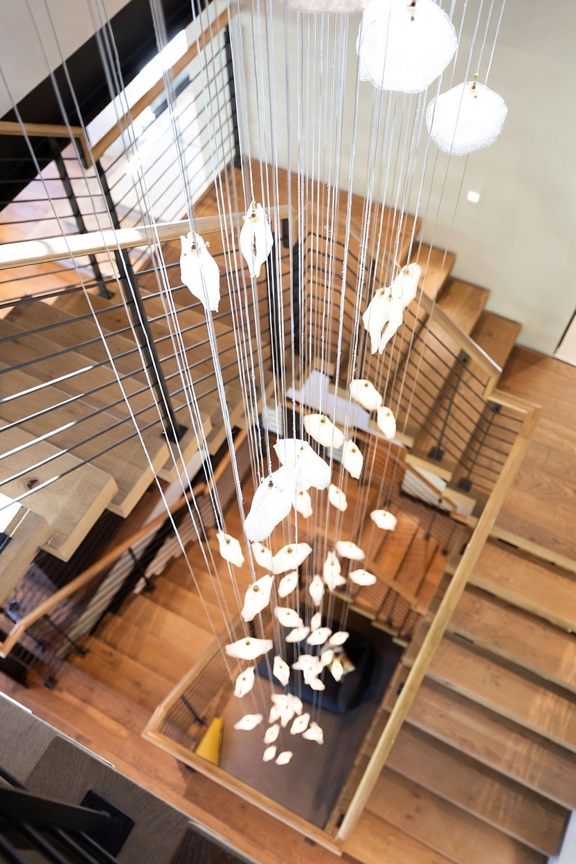 This high-angle perspective captures a dramatic, multi-level staircase featuring light wood treads and a modern metal railing system. A striking, sculptural chandelier with numerous translucent, leaf-like pendants hangs centrally through the open stairwell, creating a focal point that draws the eye downward. The clean lines and open design emphasize a contemporary architectural aesthetic.