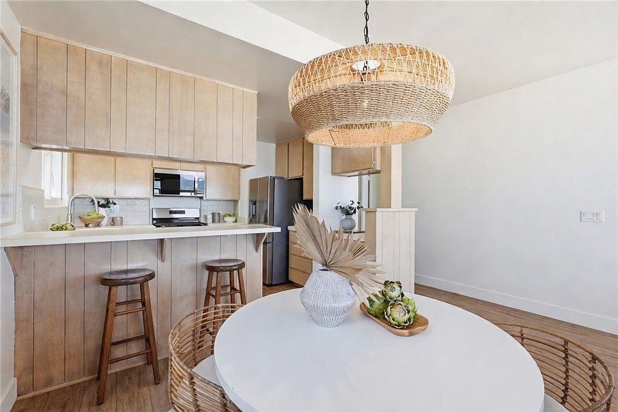 This bright, open-concept kitchen and dining area features light wood cabinetry and a breakfast bar with two wooden stools. A round white dining table sits in the foreground, illuminated by a large, textured woven pendant light fixture. The space has a clean, coastal-inspired aesthetic with light wood-look flooring and neutral walls.