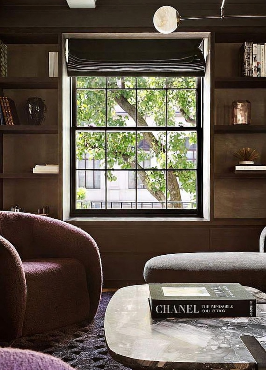 This sophisticated study features dark, built-in shelving flanking a large, multi-pane window that provides a view of lush green trees. In the foreground, a plush, textured armchair sits beside a low, stone-topped coffee table holding a Chanel coffee table book. The room is illuminated by a modern, minimalist light fixture, creating an intimate and refined atmosphere.