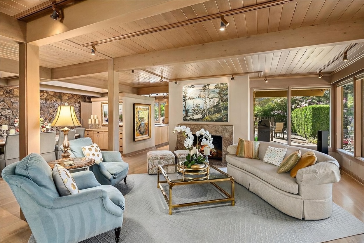 This spacious living room features a warm, wood-paneled ceiling with exposed beams and recessed lighting. The room is anchored by a fireplace with a stone-clad chimney breast and a glass-topped coffee table, surrounded by plush seating including a curved sofa and two armchairs. Large sliding glass doors provide a seamless connection to the outdoor patio, filling the space with natural light and offering a view of the lush greenery outside.